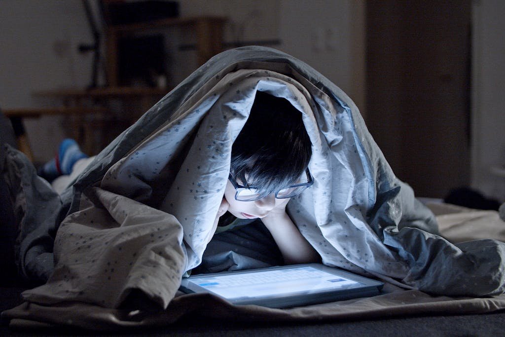A child with glasses watching a tablet under a blanket in a dimly lit room. Nighttime setting with a cozy atmosphere.